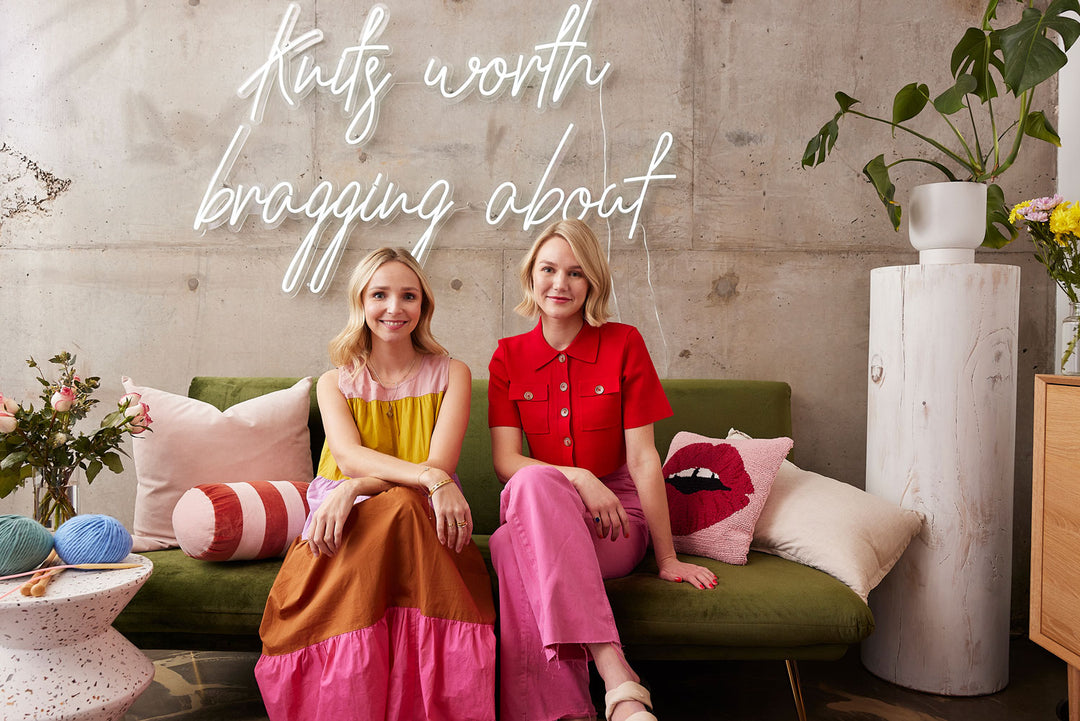 Cardigang co-founders Morgan and Cat sitting on green couch, smiling at camera. They are sitting in front of a neon sign that says "Knits Worth Bragging About"