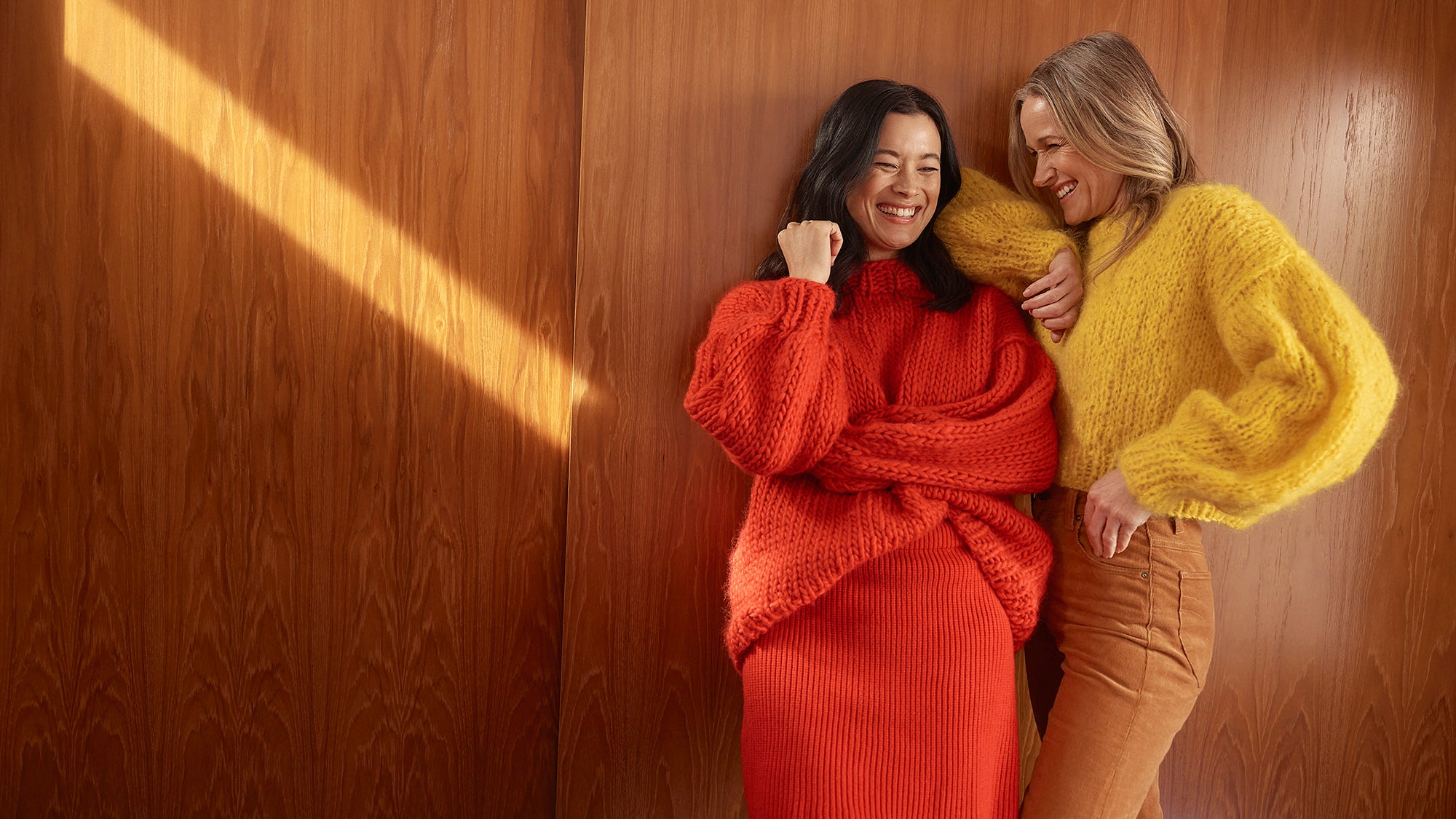 Two women are laughing in front of a wooden door. They are both wearing bright colourful fluffy chunky knitted jumpers.