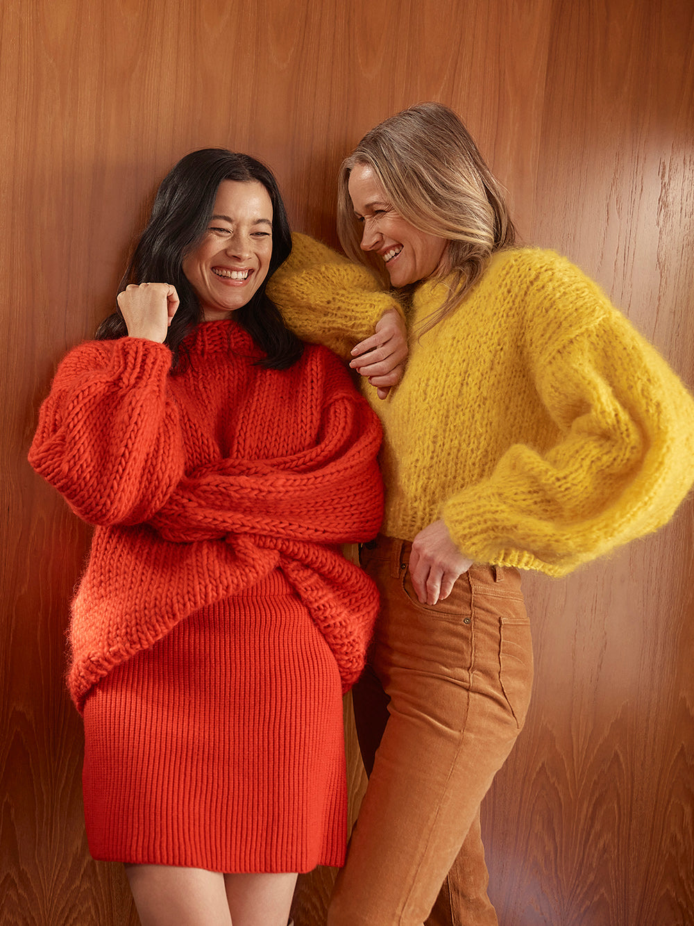 Two women are laughing in front of a wooden door. They are both wearing bright colourful fluffy chunky knitted jumpers.