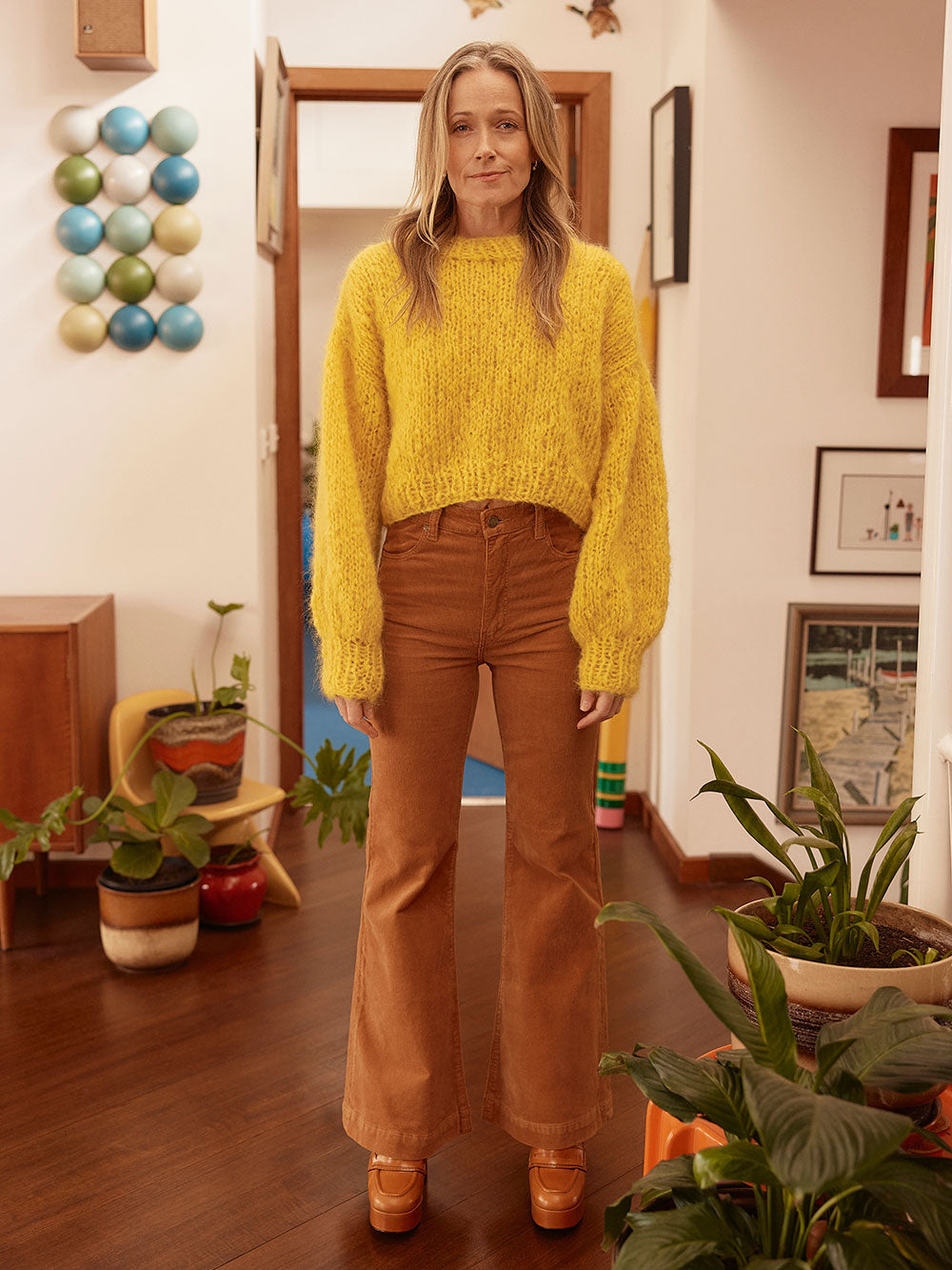 Woman stands in hallway wearing a bright yellow chunky knitted mohair jumper and brown cord jeans. 