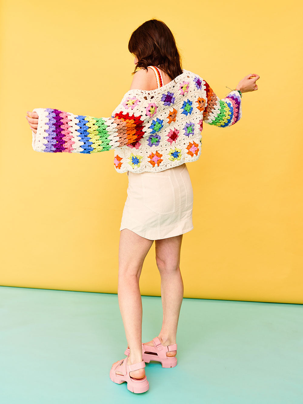 Woman with dark wavy hair stands with her back facing the camera, she is wearing a colourful granny square crocheted cardigan from Cardigang.
