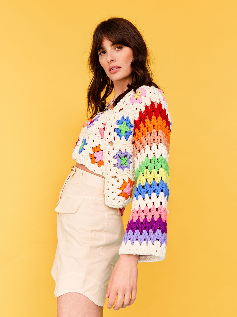 Woman with dark wavy hair stands side on the the camera, she iswears a colourful granny square crocheted cardigan from Cardigang.