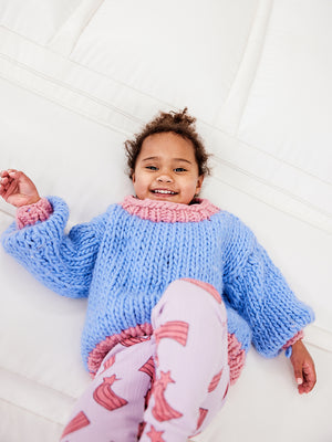 A girl is smiling lying down on a white jumping castle. She is wearing a blue chunky knitted jumper and purple and pink leggings