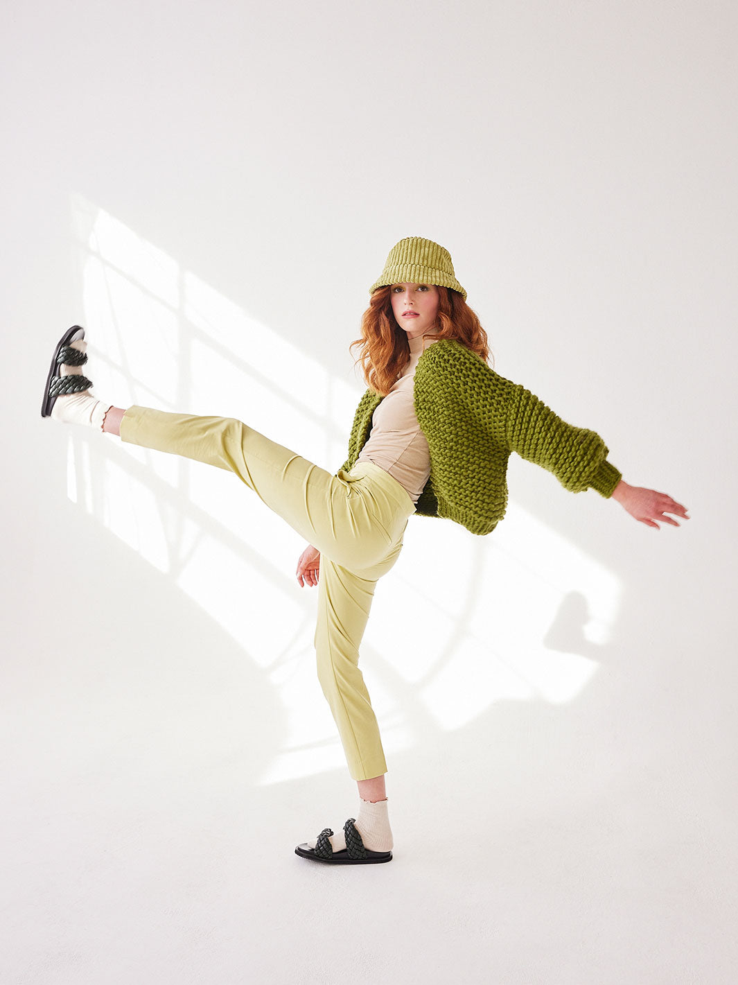 Girl in hat kicking leg to the sky, wearing a green chunky knitted cardigan