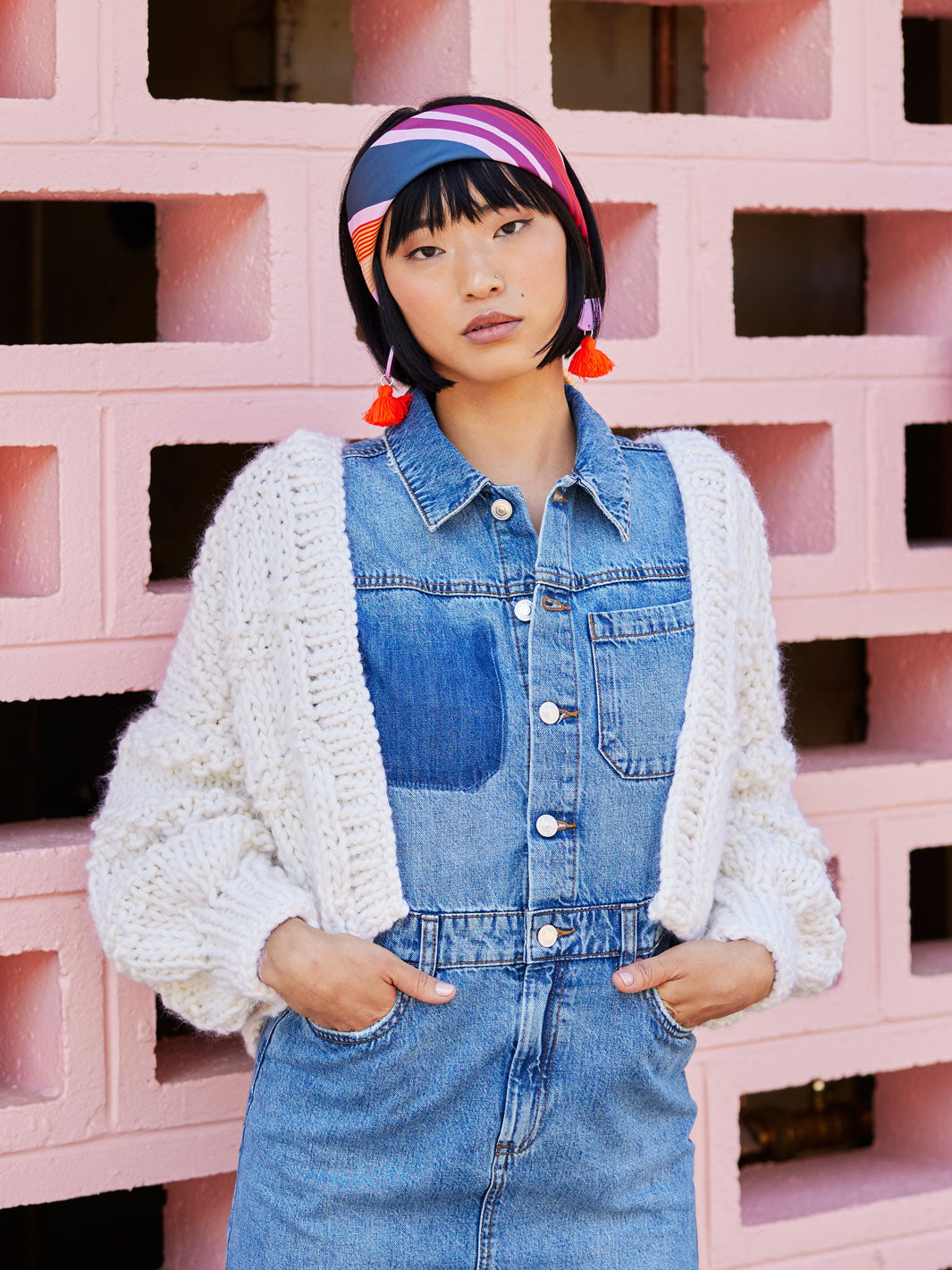Woman standing in front of pink wall waring a denim dress and white chunky knitted cardigan