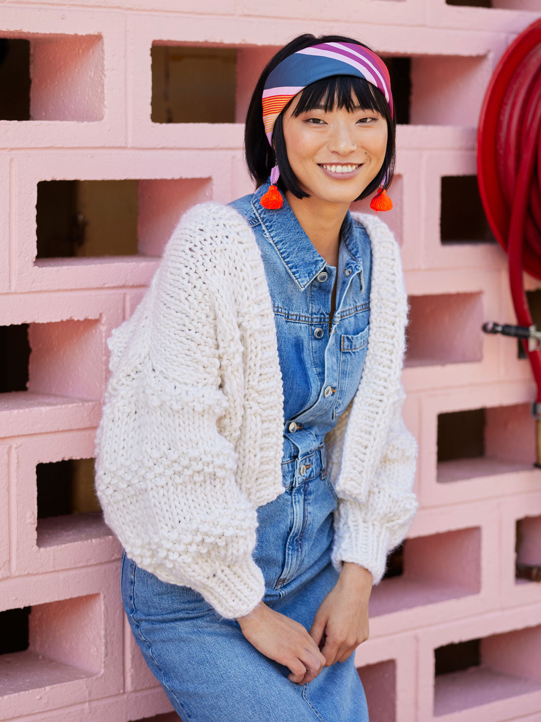 Woman leans on pink wall wearing a white chunky knitted cardigan