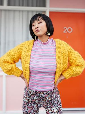 Woman in motel carpark stands with hands on hips and wears a bright yellow chunky knitted cardigan