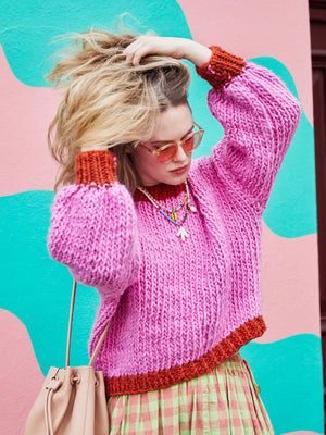 Playing with hair in front of colourful wall wearing bright chunky knit Betty jumper