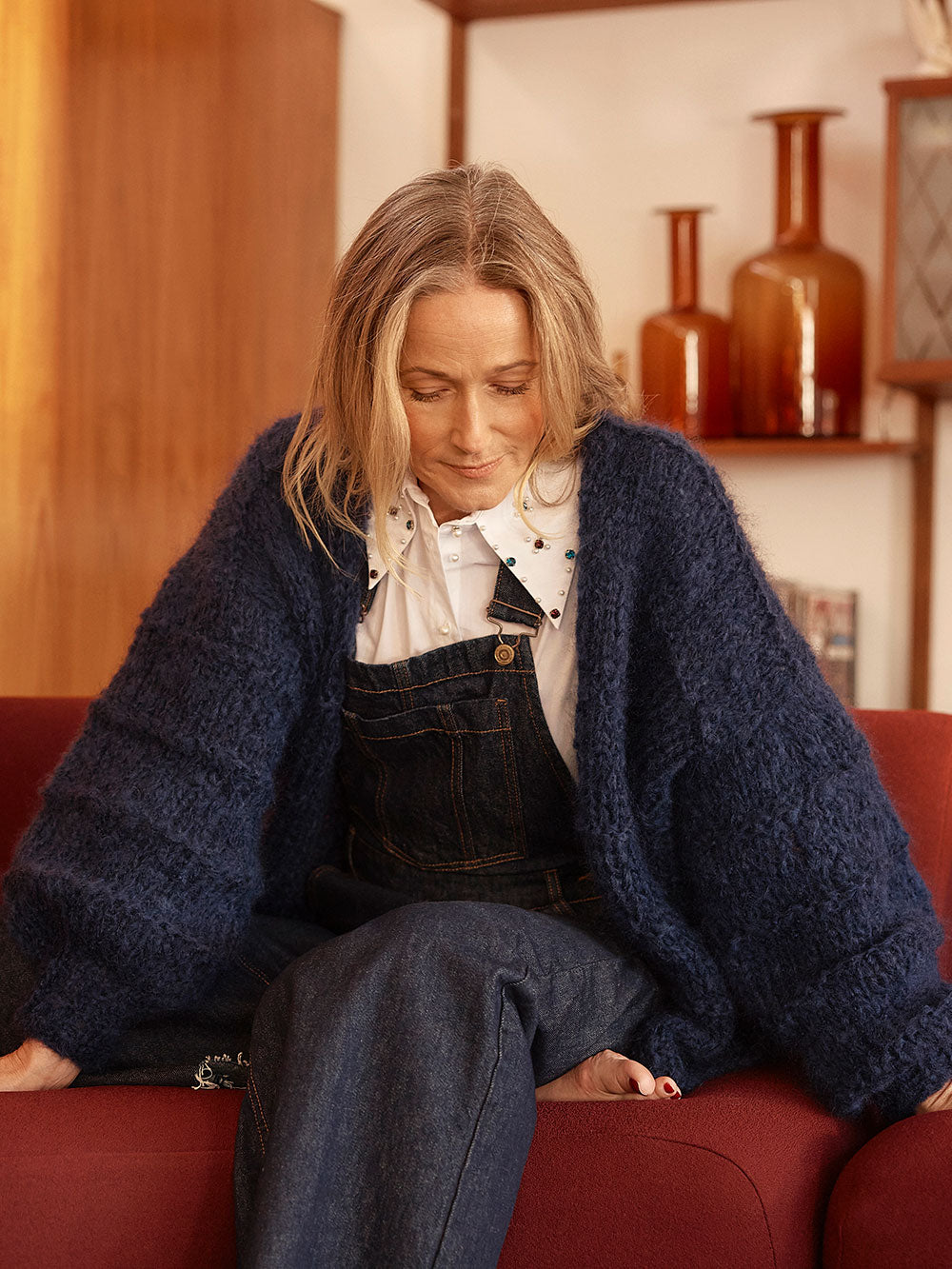 Woman sits on red couch, she is smiling at the camera and wearing a navy blue knitted mohair cardigan and denim overalls with a white collared shirt.
