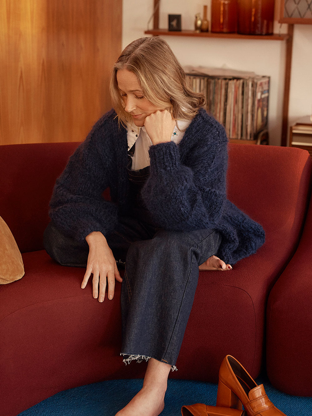 Woman sits on red couch, she is smiling at the camera and wearing a navy blue knitted mohair cardigan and denim overalls with a white collared shirt.