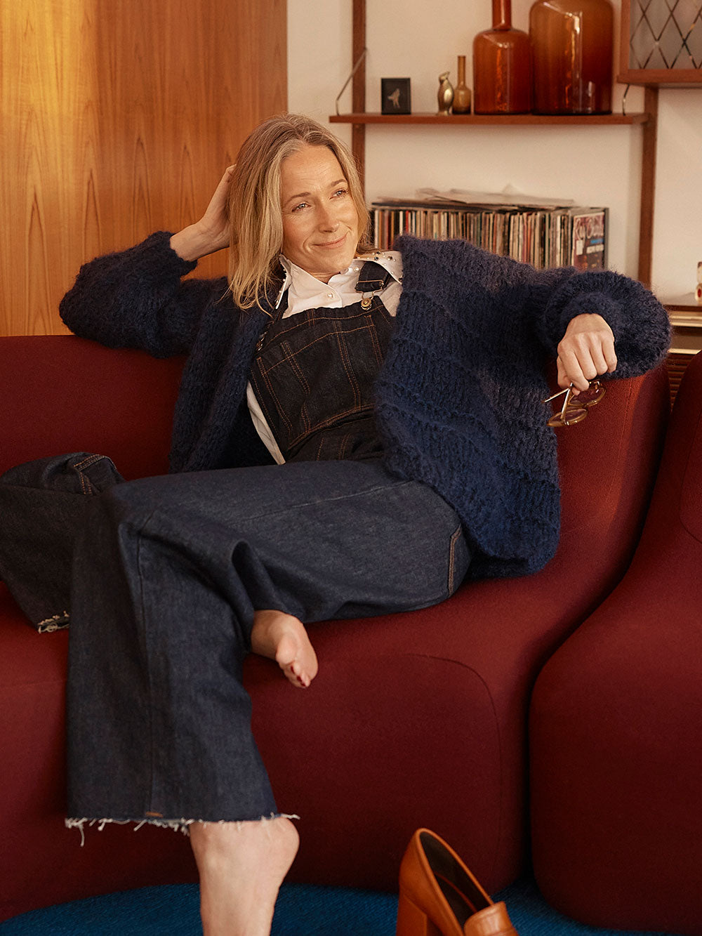 Woman lounges on red couch, she is smiling at the camera and wearing a navy blue knitted mohair cardigan and denim overalls with a white collared shirt.