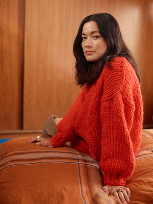 Woman sits on a bed wearing a bright red chunky knitted jumper and red skirt