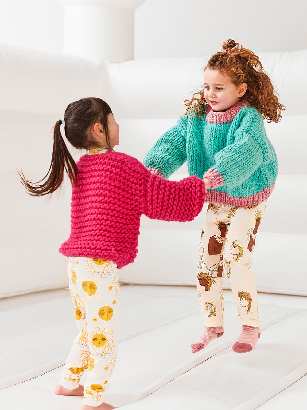 Two girls are jumping on a white jumping castle, they are both wearing chunky knitted jumpers and cardigans