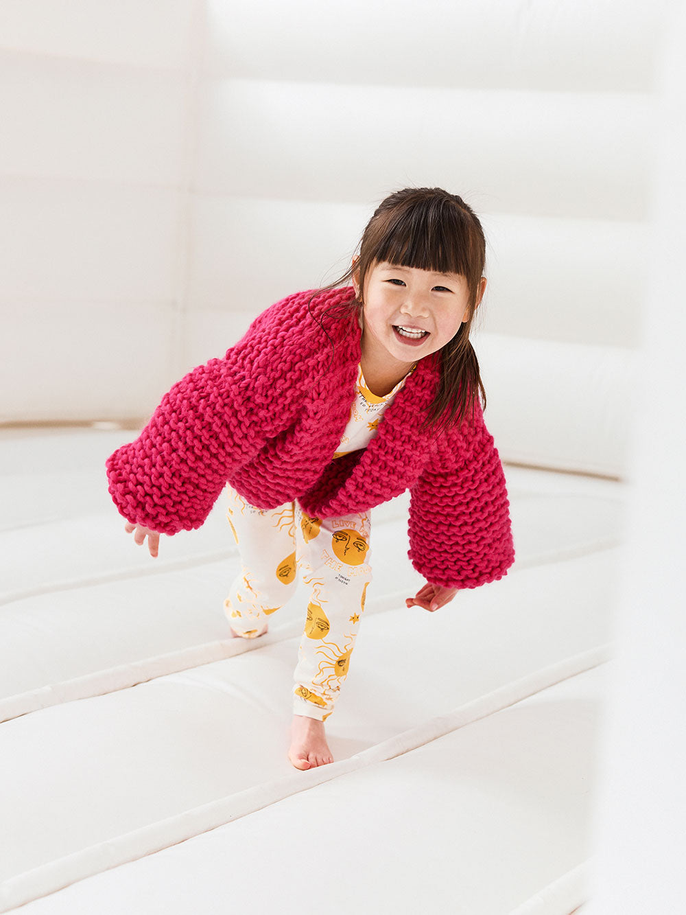 Little girl runs on a white jumping castle smiling at the camera and wearing a bright pink chunky knitted cardigan and leggings