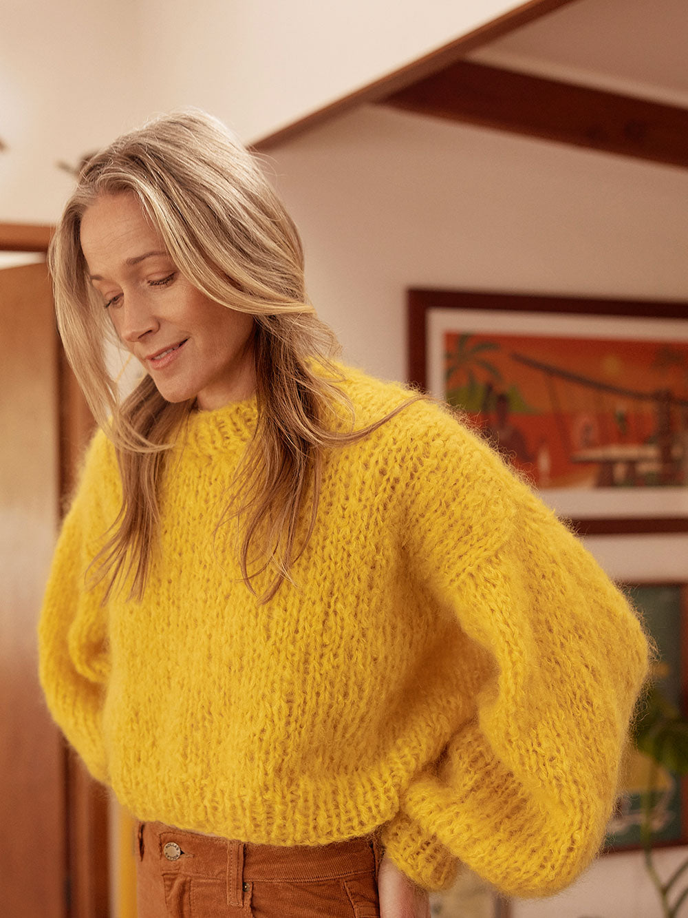Woman smiles at camera wearing a bright yellow chunky knitted mohair jumper and brown cord jeans. 
