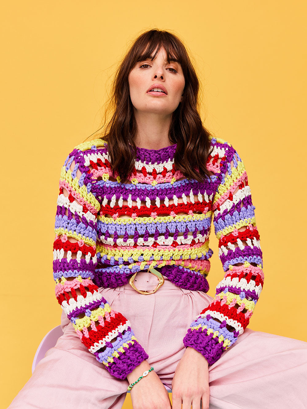 Woman with dark wavy hair is sitting on a lilac stool wearing a DIY striped crochet jumper by Cardigang