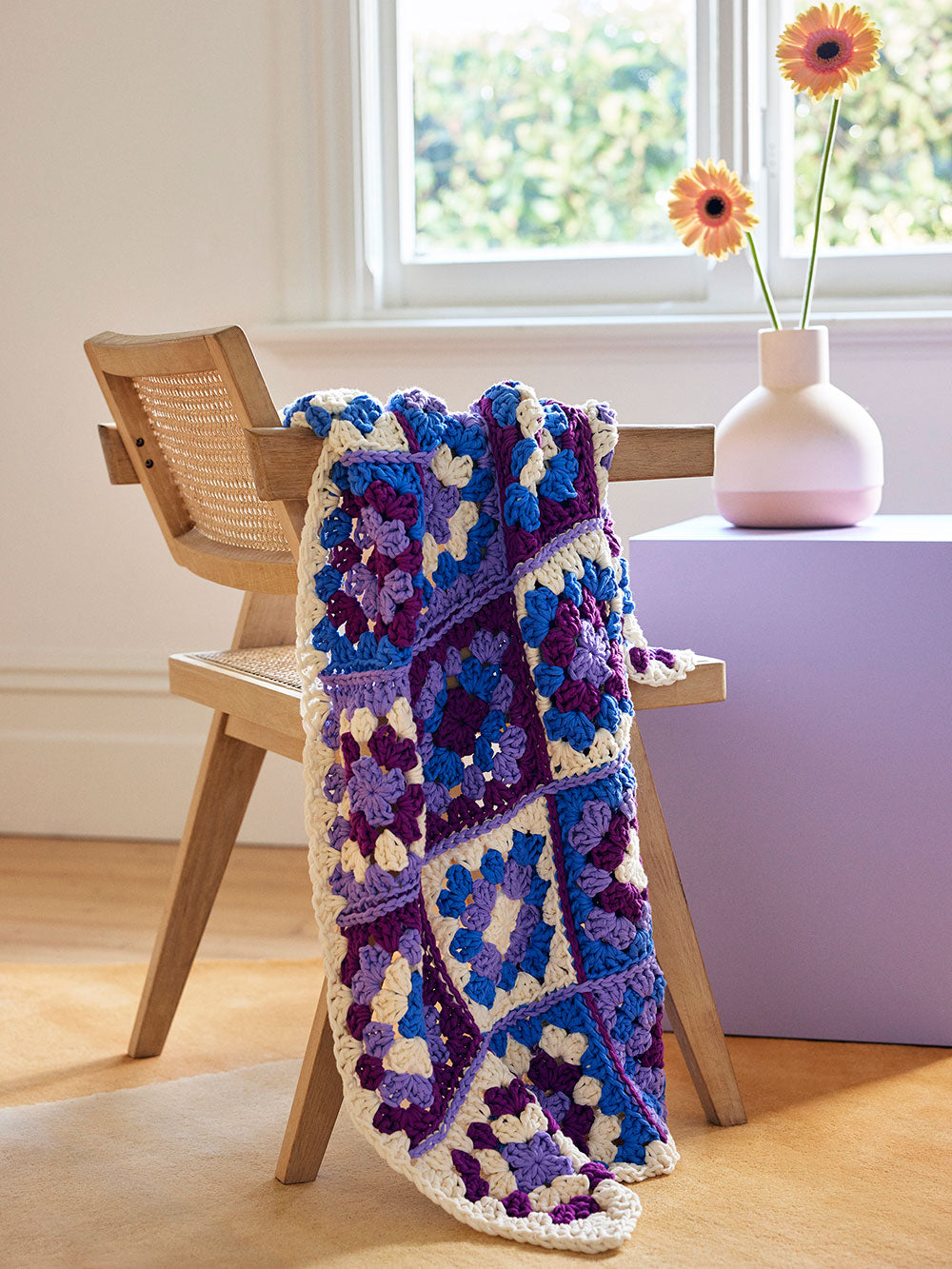 Image of a multi-colour granny square baby blanket draped over the side of a rattan chair,