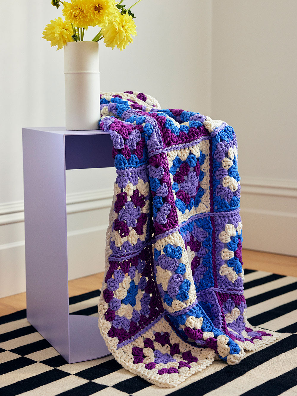 Image of a multi-colour granny square baby blanket draped over a purple side table with a vase of yellow flowers. 