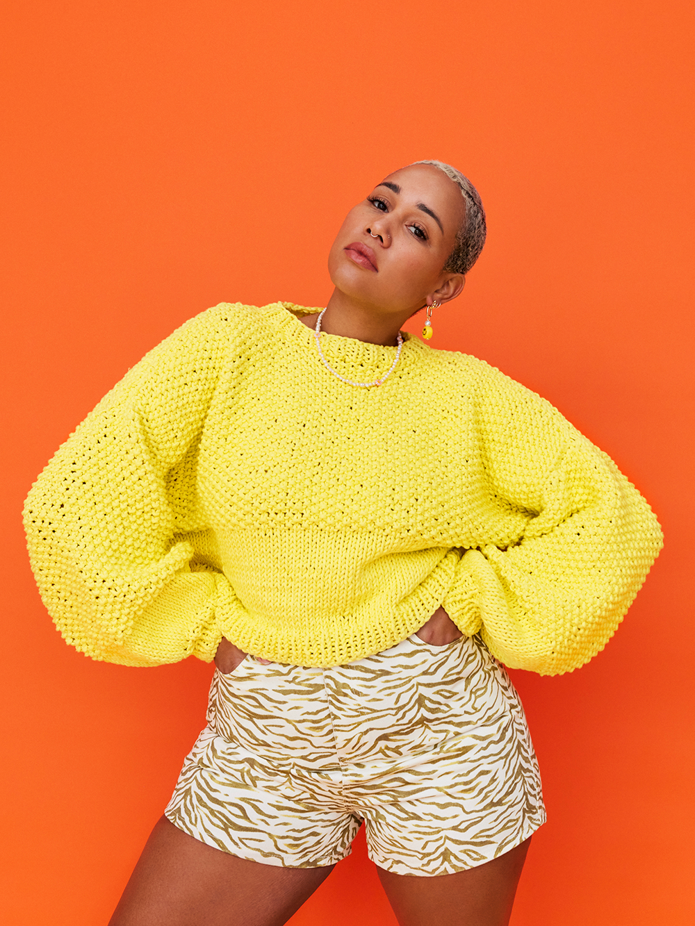 Woman stands in front of orange background, wearing a chunky knitted oversized yellow cotton jumper and patterned denim shorts.