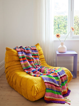 Rainbow coloured Florence afghan crocheted blanket is draped over a yellow suede chair