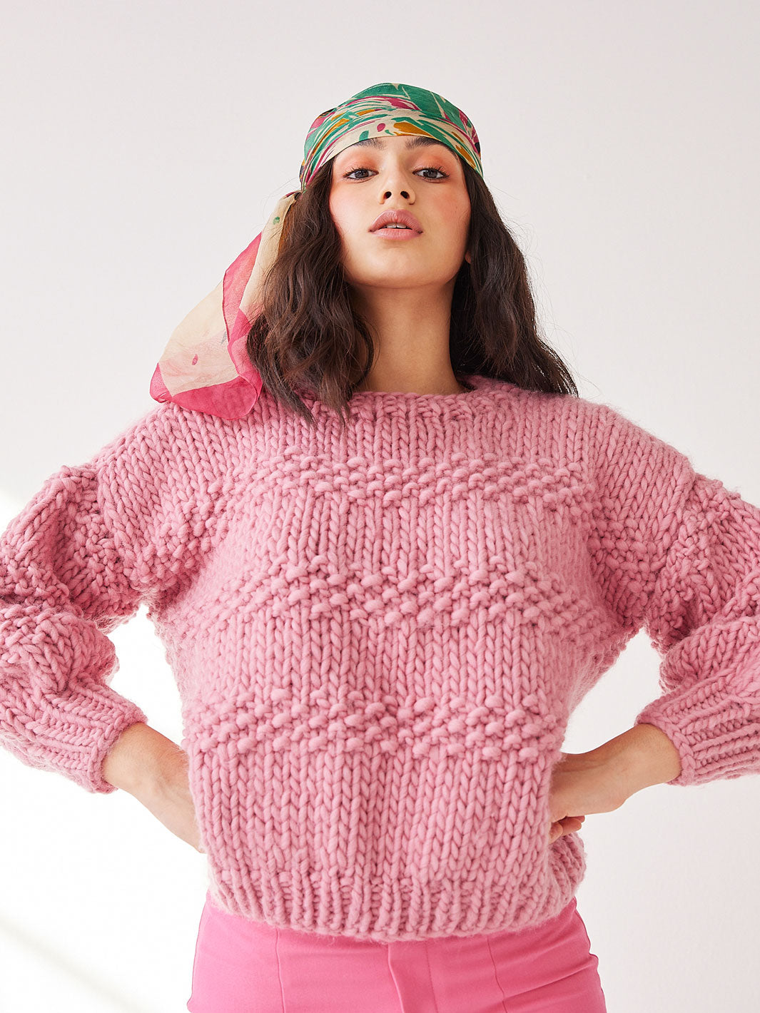 Woman stands with hands on hips. She is wearing a chunky knitted pink jumper, pink pants and a scarf on her head