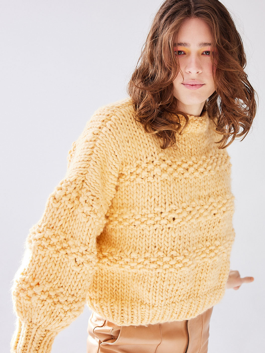 Woman looks directly at camera, she is wearing a chunky knitted yellow jumper and brown leather pants