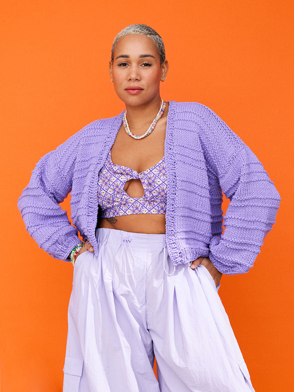 Woman standing in front of orange backdrop wears a lilac coloured, chunky knitted cotton cardigan, a purple bathers top and lilac coloured cargo pants.
