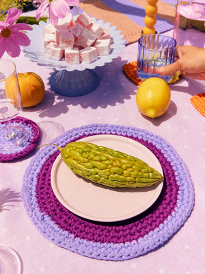 Close up of Isla plum and lilac circular crochet placemats and coasters styled on a table with vegetables and glassware. 