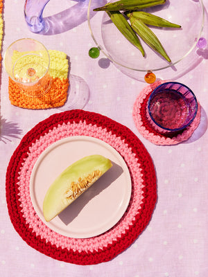 Isla red and pink circular crochet placemats and coasters styled on a table with vegetables and glassware. 