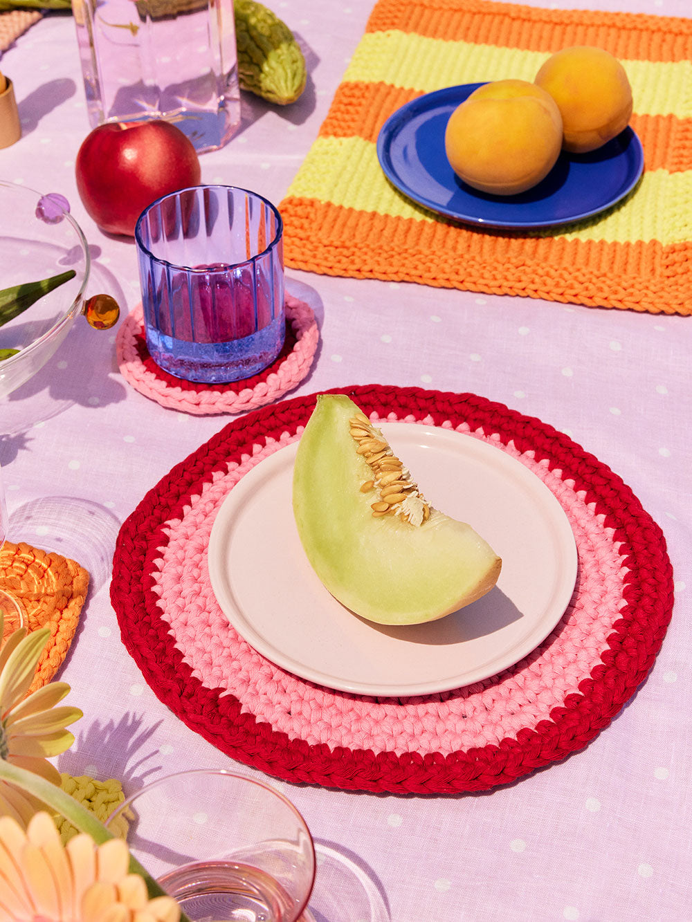 Isla red and pink circular crochet placemats and coasters styled on a table with vegetables and glassware. 