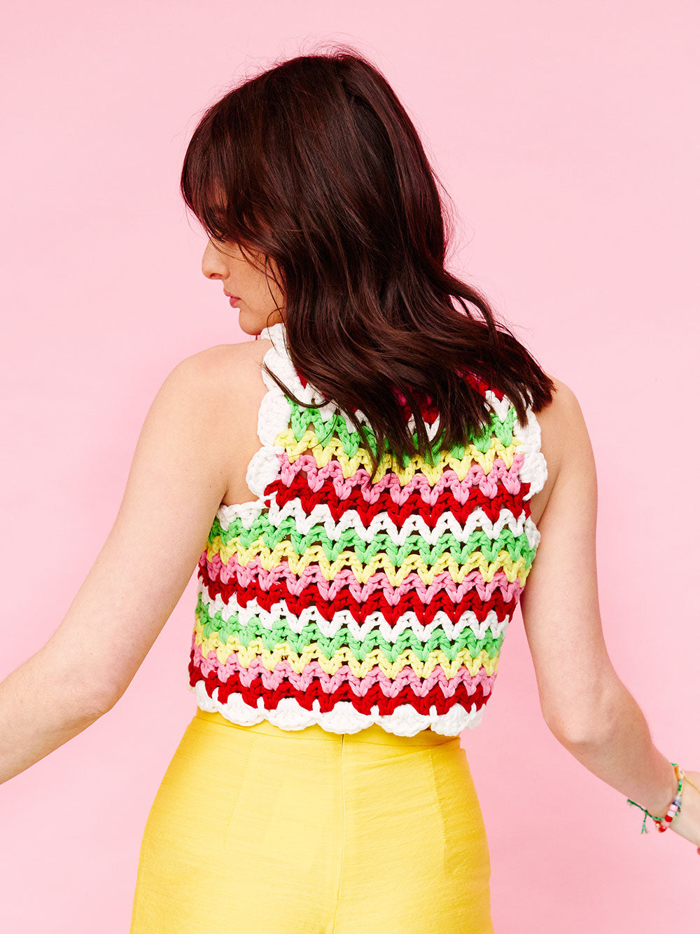 Woman with dark wavy hair has her back to the camera, she is wearing a colourful, crocheted cotton stripe tank top and yellow pants. 