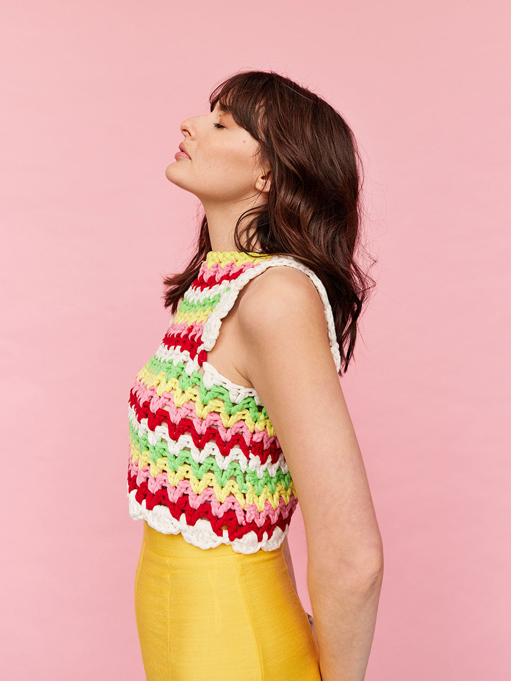 Woman with dark wavy hair is side on to the camera, she is wearing a colourful, crocheted cotton stripe tank top and yellow pants.
