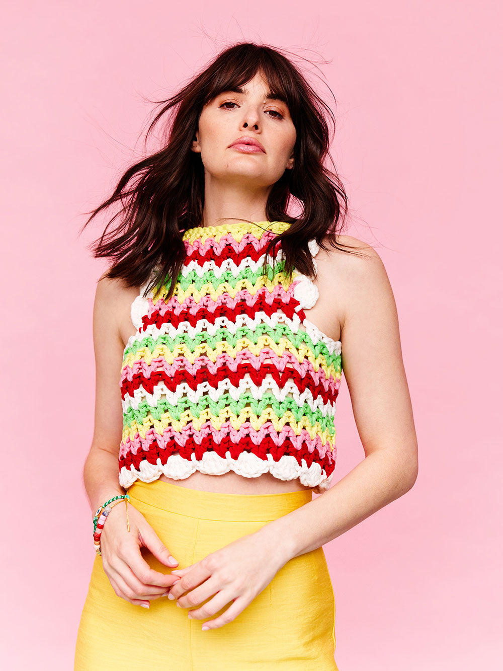 Woman with dark wavy hair is wearing a colourful, crocheted cotton stripe tank top and yellow pants. 