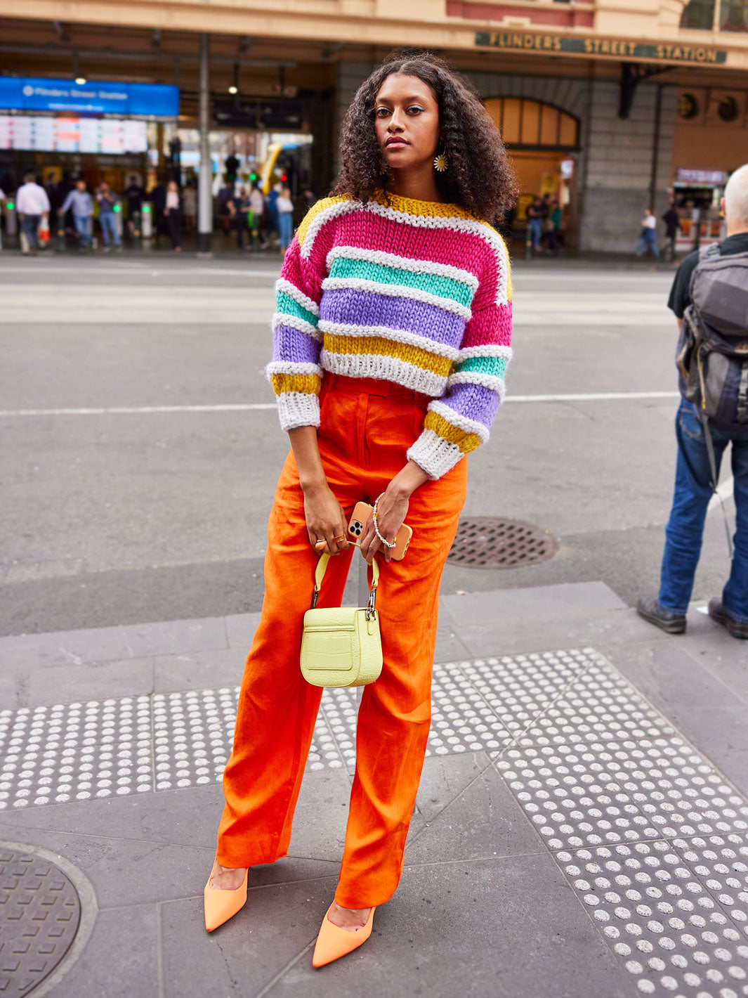 Woman leans on pole at road crossing holding a yellow handbag and wearing orange pants and a brightly coloured hand knitted jumper
