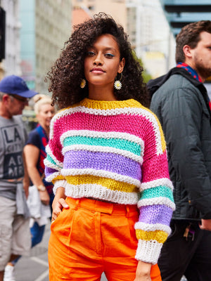 Woman stands in crowd with hand in pocket smiling at camera wearing a bright coloured striped chunky knit jumper