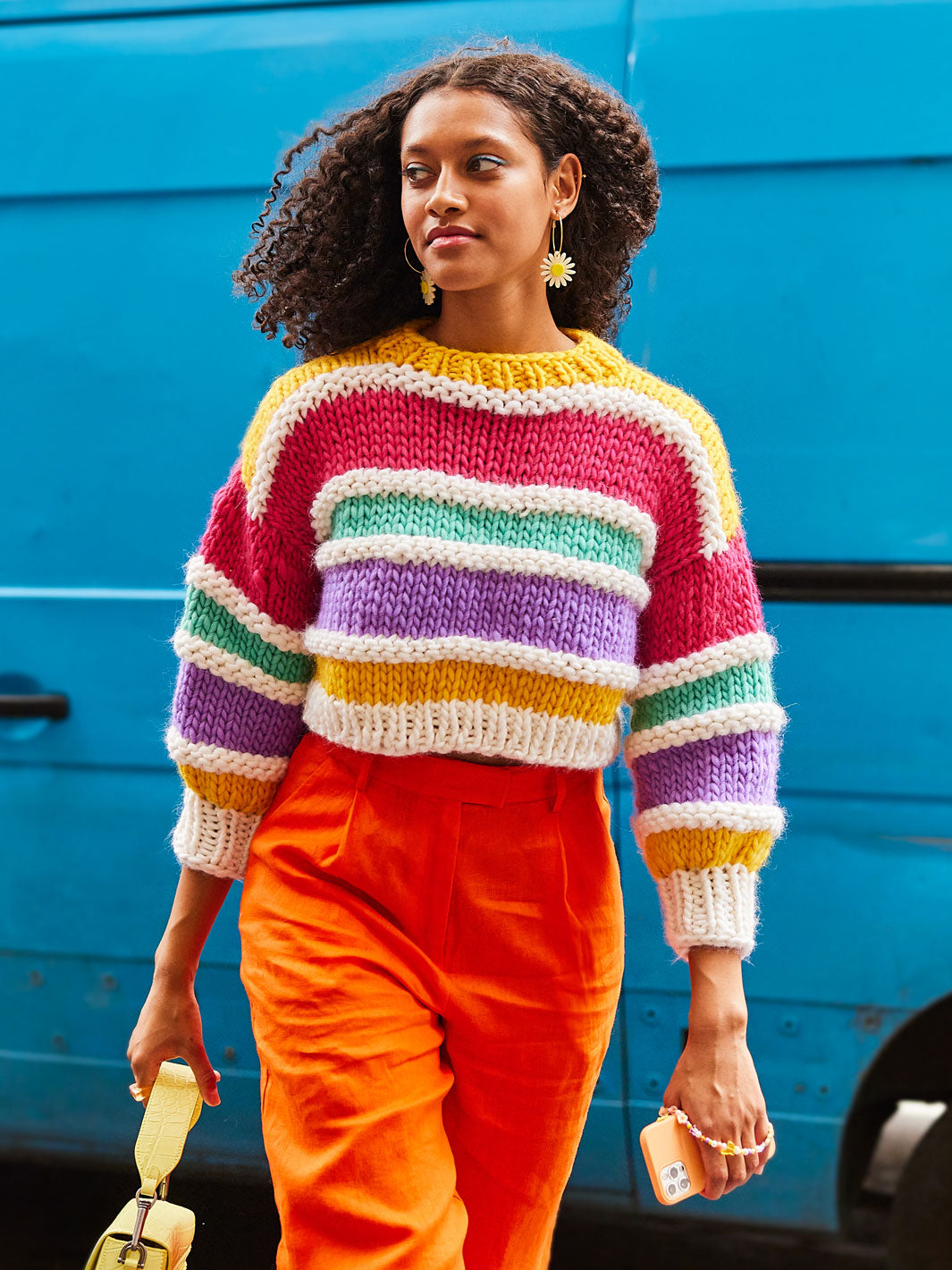 Woman walks away from blue van wearing bright orange pants and a colourful chunky knit jumper