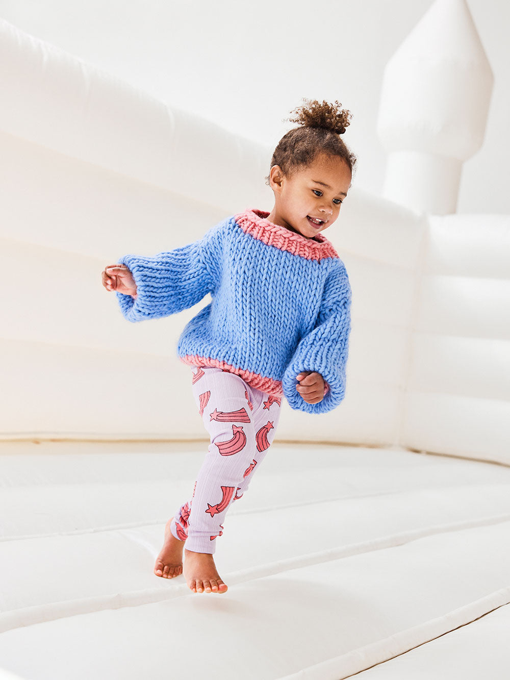 Little girls runs on white jumping castle wearing a pink and blue chunky knitted jumper and purple leggings