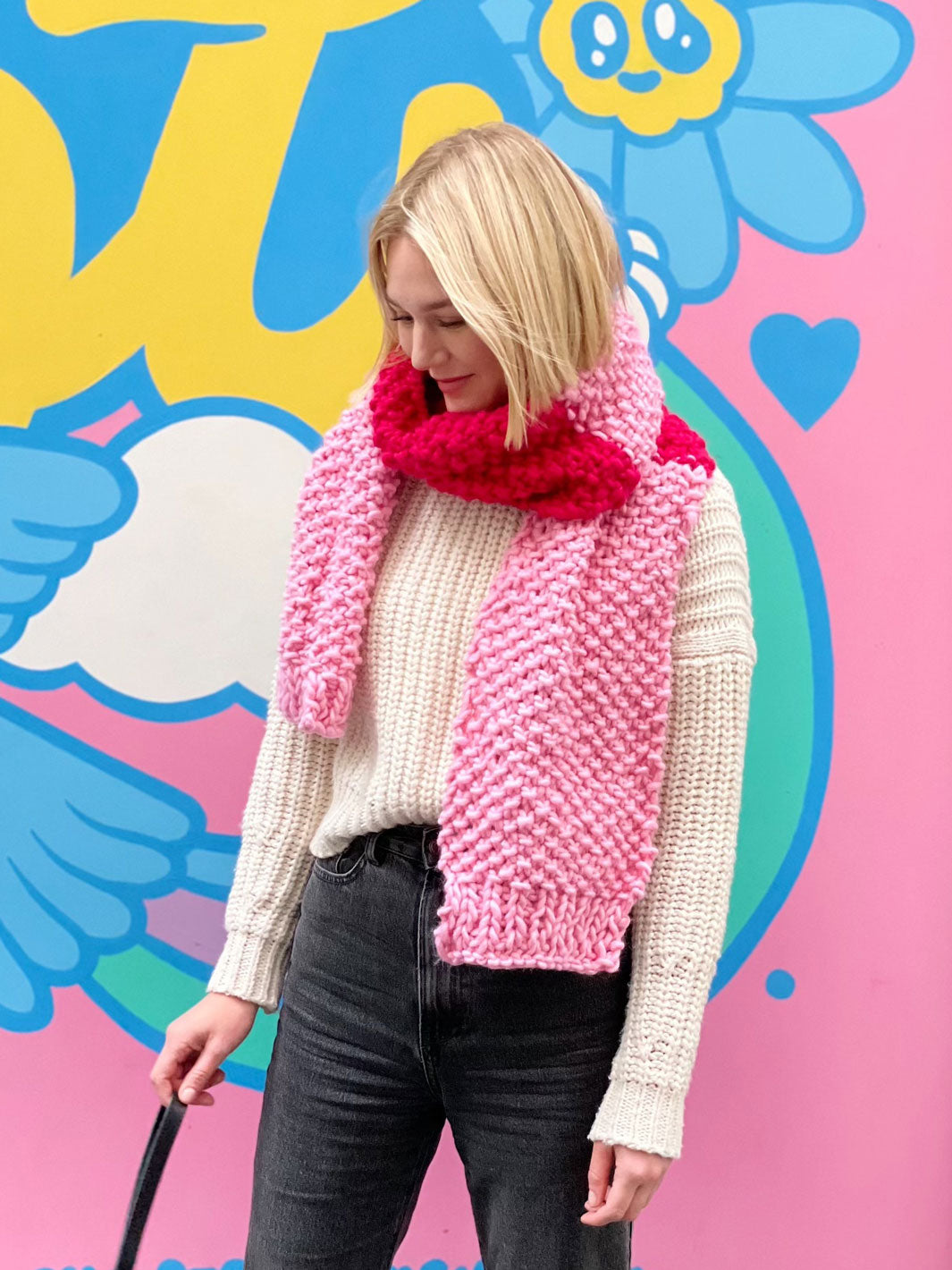 Womans stands in front of colourful wall wearing a pastel and bright pink chunky knitted scarf