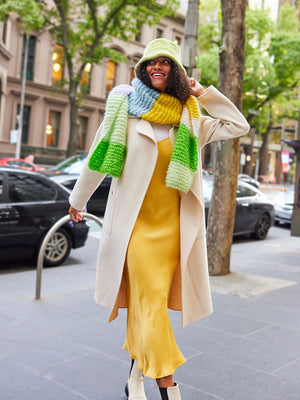Woman in yellow dress wears a chunky knitted colourful scarf wrapped around her neck and a bucket hat